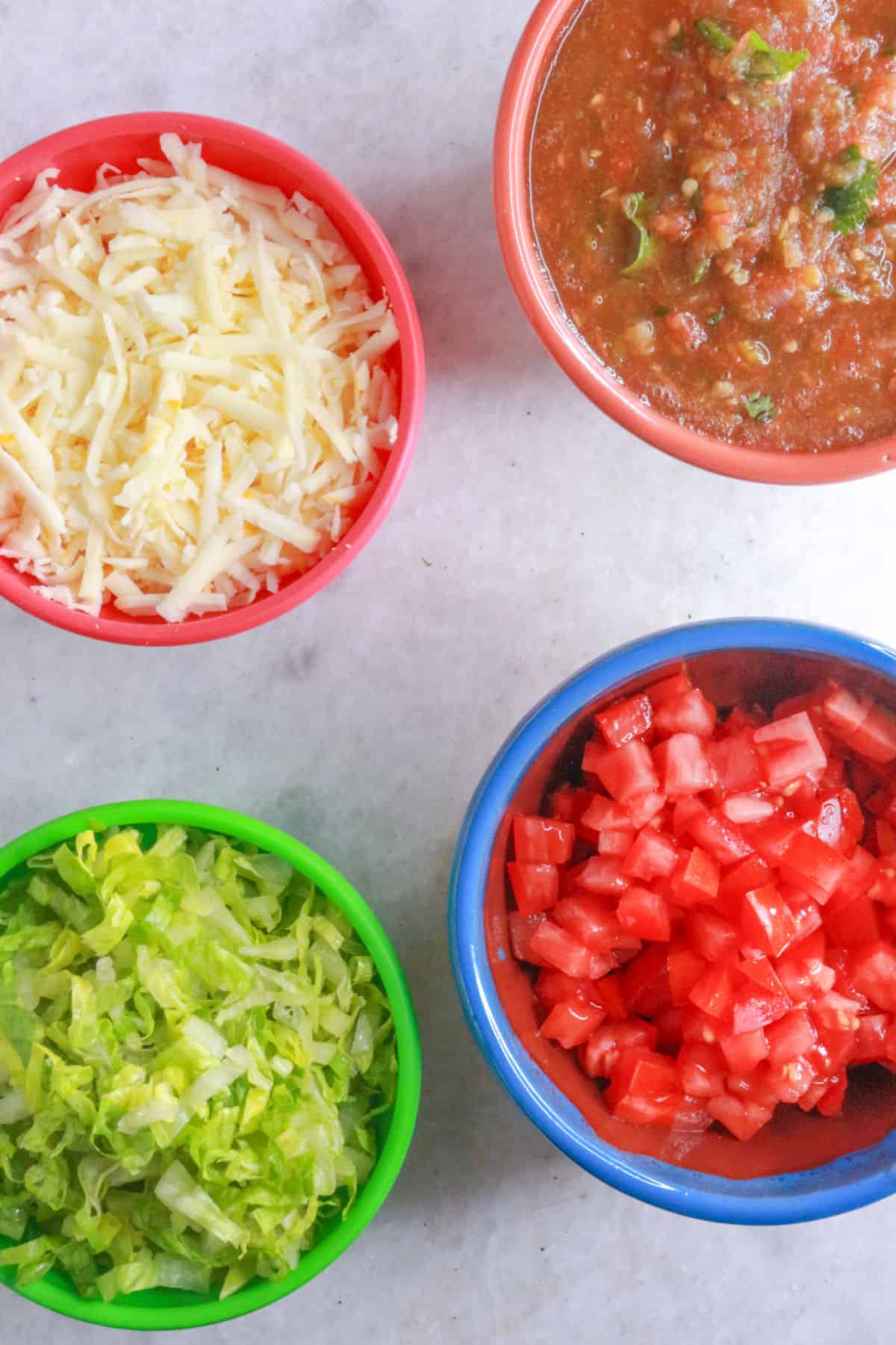A bowl with shredded cheese, a bowl with jalapeño salsa, a bowl with shredded lettuce, a bowl with diced tomato