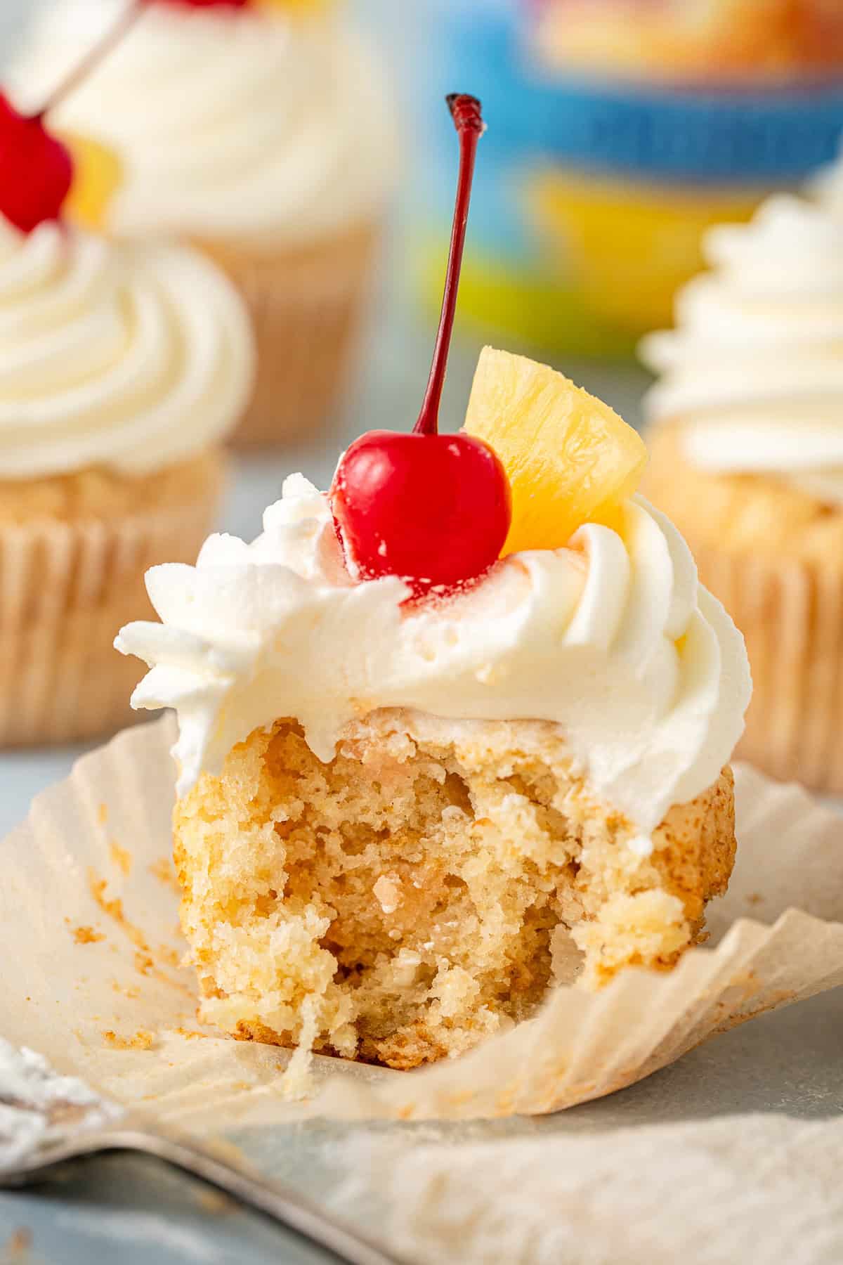A close up of a vegan pineapple cupcake with a bite taken and cherry on top.