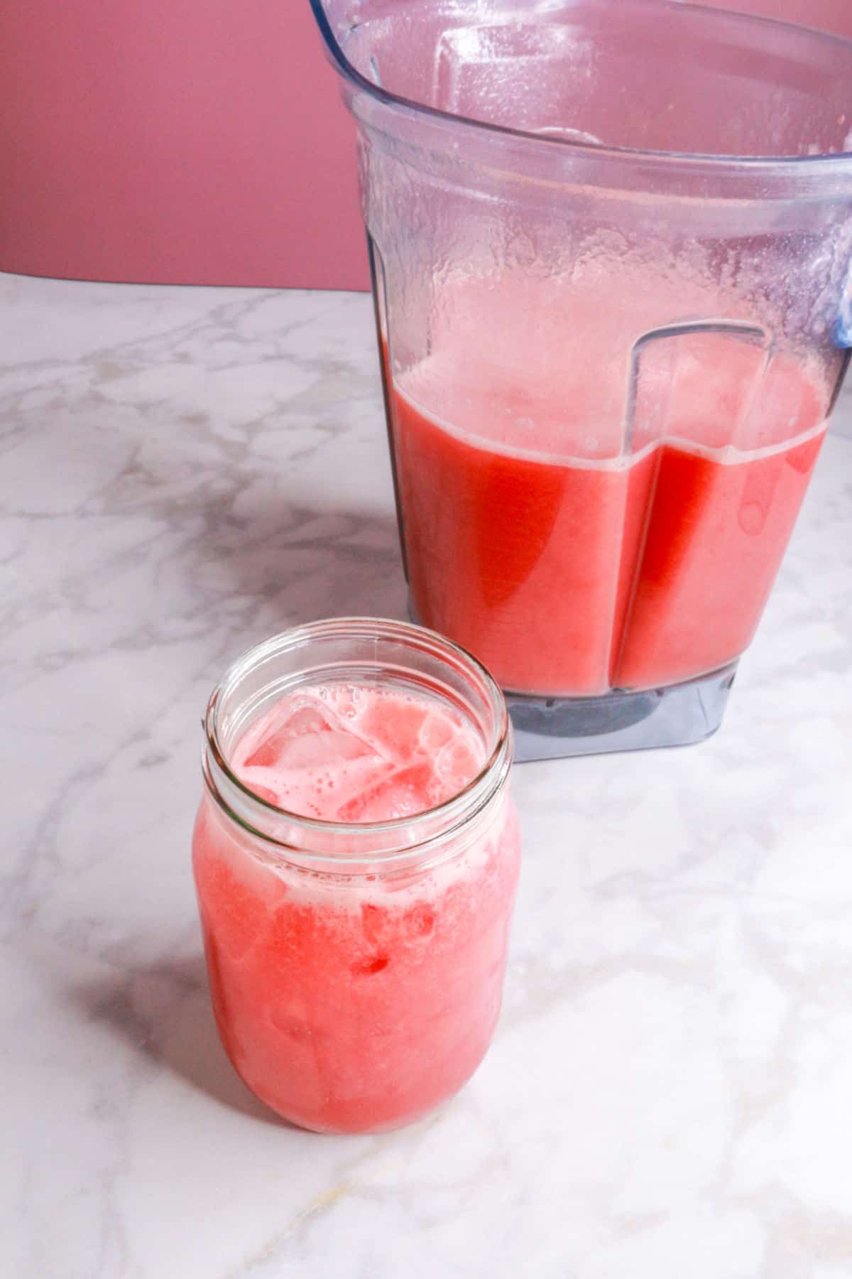 A glass of watermelon strawberry agua fresca in a glass with a blender behind it