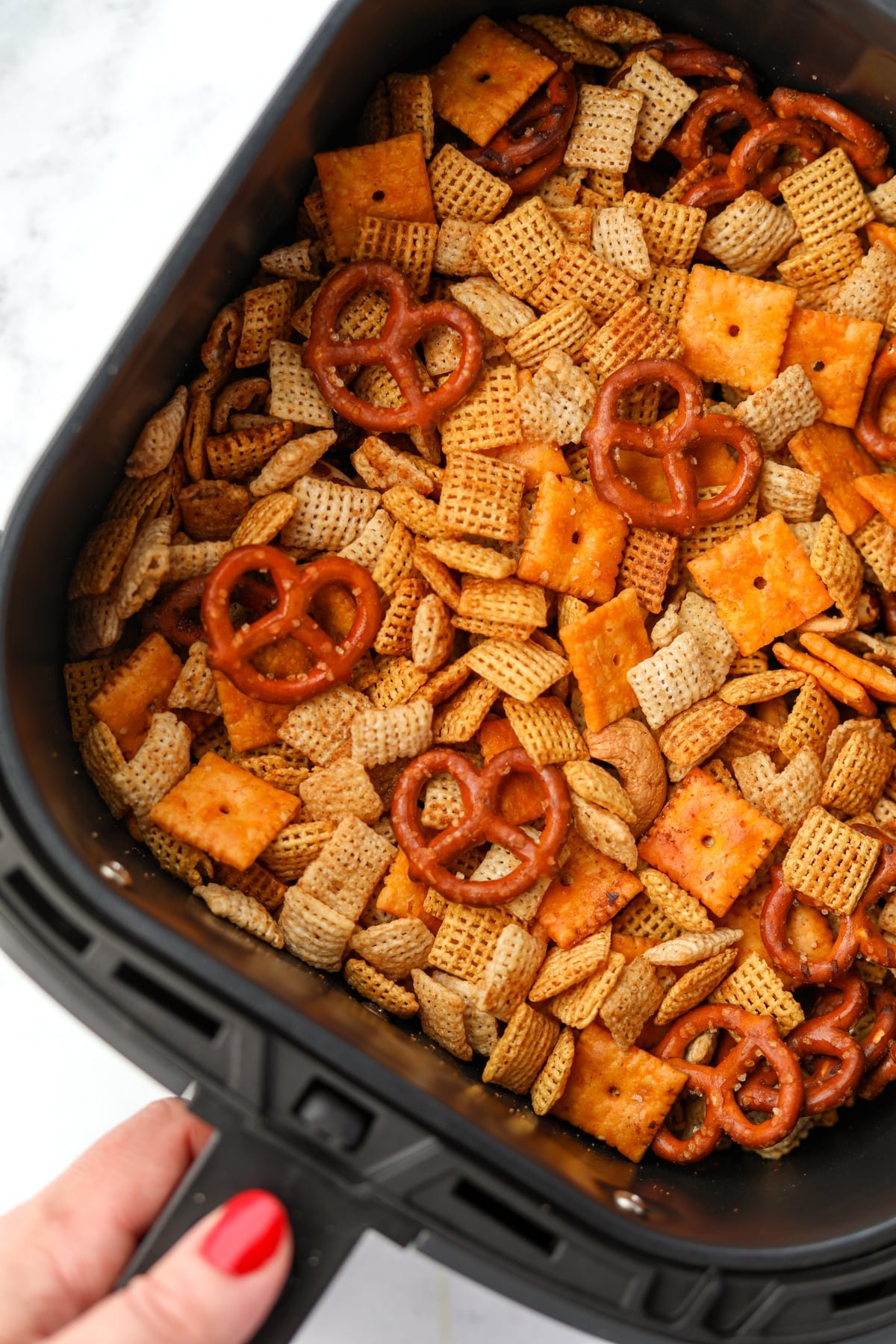 A hand removing an air fryer basket filled with a homemade snack mix.