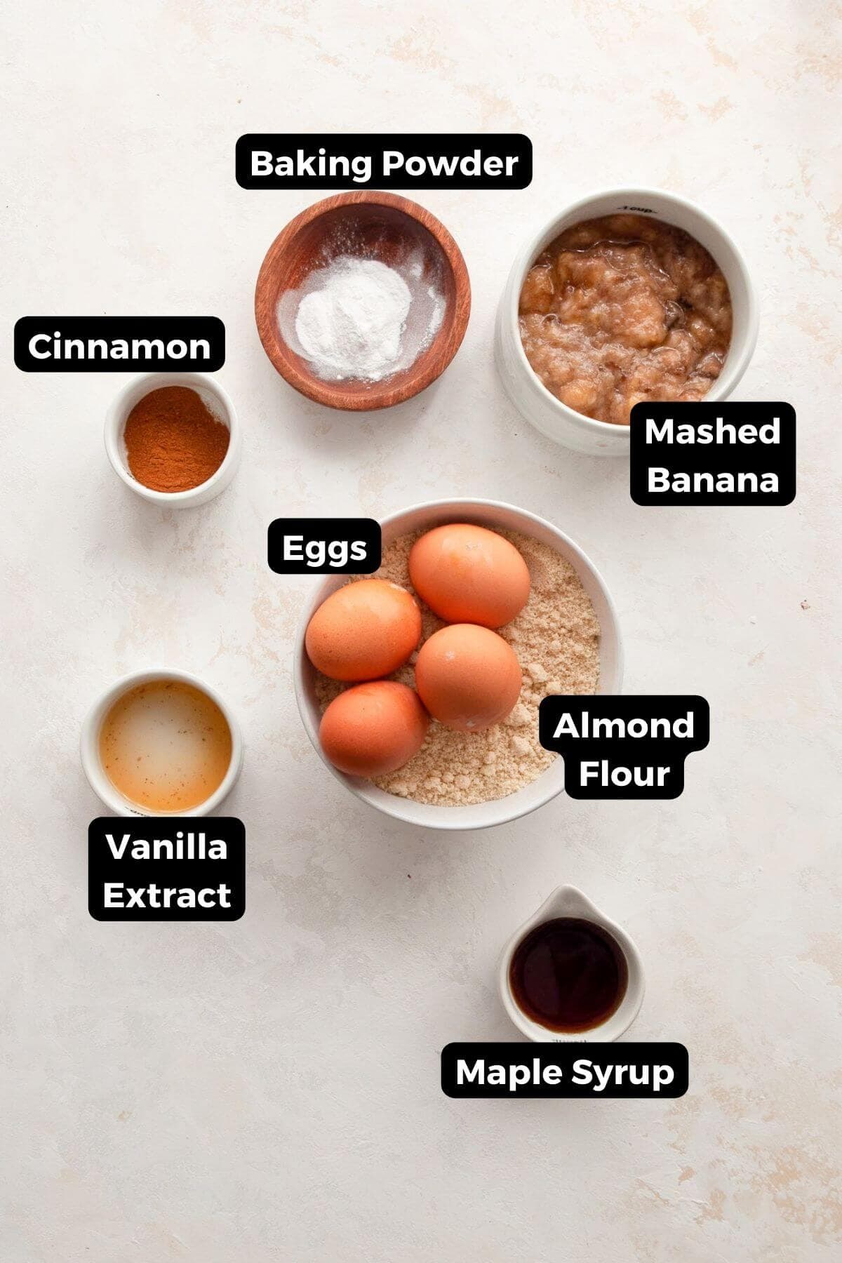 All ingredients for almond flour banana pancakes arranged on a counter: almond flour, ripe bananas, cinnamon, baking powder, eggs, vanilla extract, and maple syrup.