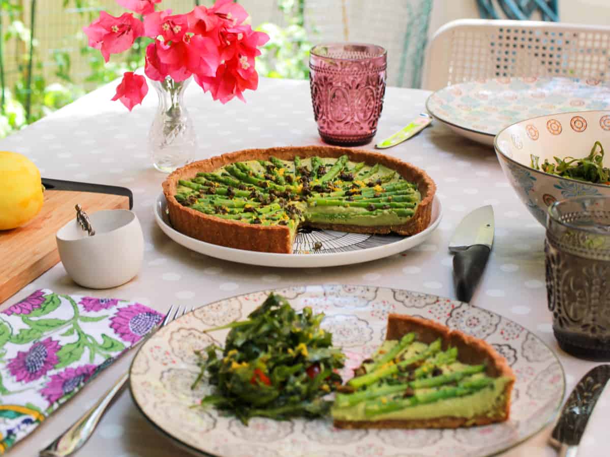 a table set for lunch in the garden with flowers on the table and an asparagus tart in the middle with a portion of tart and salad served on the nearest plate.