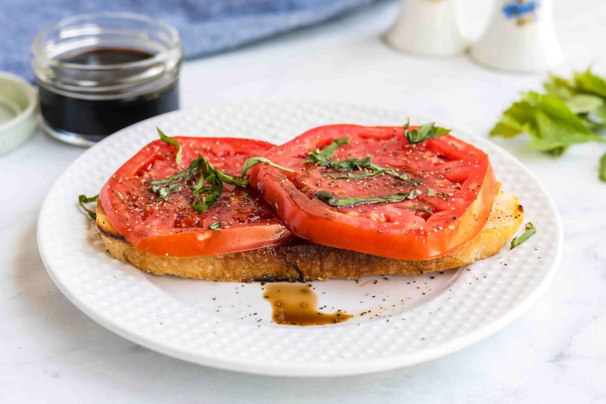 Two slices of tomato on toasted bread, garnished with chopped basil and pepper, on a white plate with some balsamic glaze drizzled nearby.