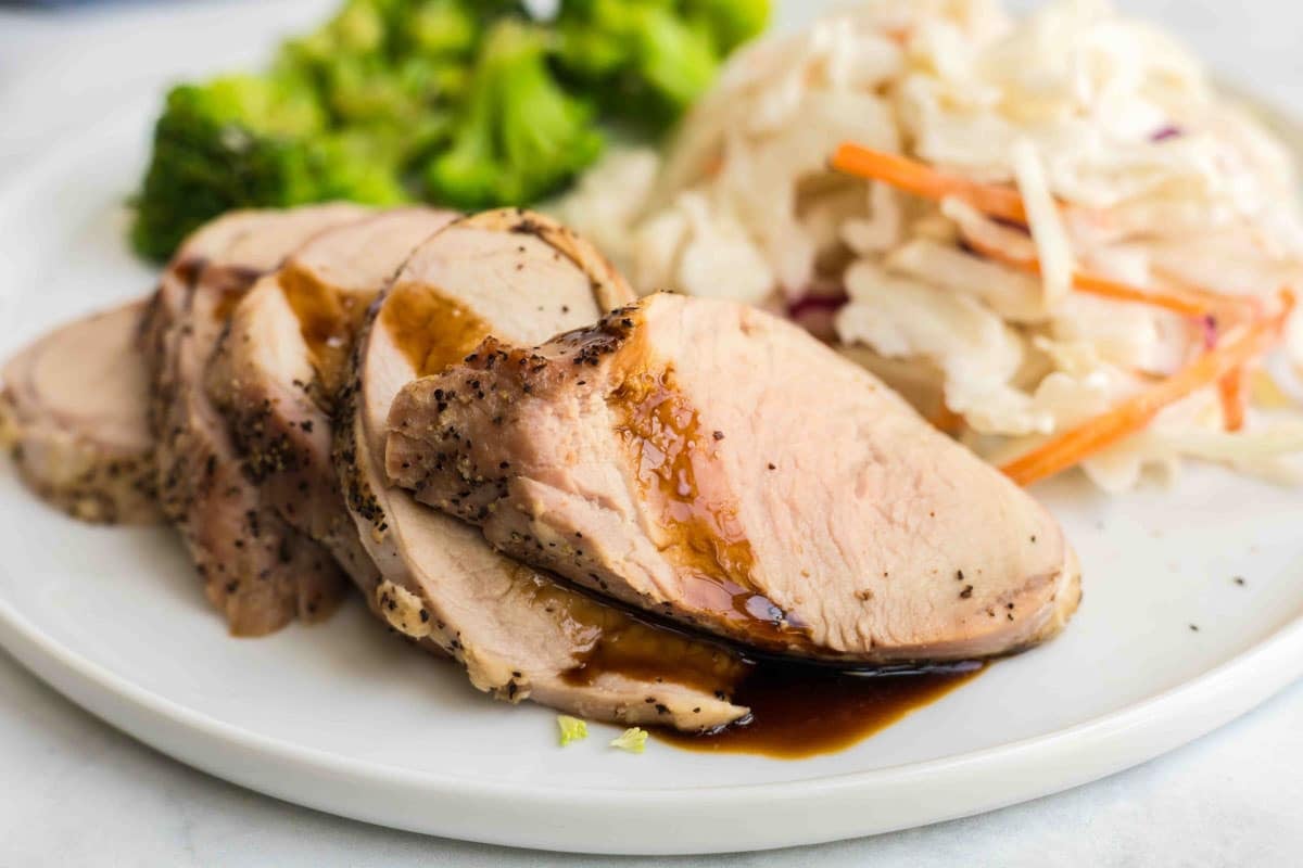 Sliced pork tenderloin with balsamic glaze served alongside broccoli and shredded coleslaw on a white plate.