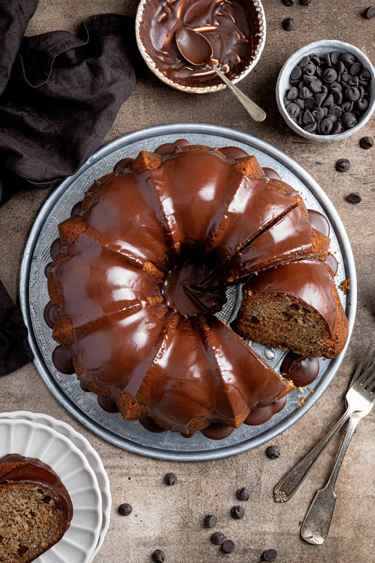 Vegan banana bundt cake on a metal plate with a couple of slices removed surrounded by bowls of chocolate chips and chocolate glaze.