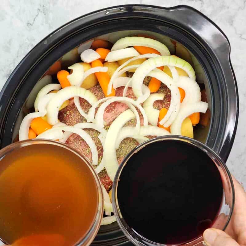 Overhead view of process shot 3 - add the sliced onion, potato halves, and carrots around and on top of the roast and pour the red wine and beef broth into the slow cooker.