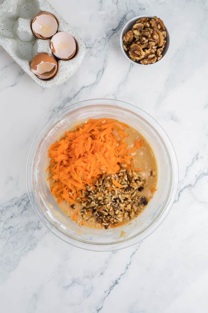 A glass bowl with cake batter, shredded carrots, and chopped walnuts sits on a marble countertop. Next to it are cracked egg shells in a dish and a small bowl of walnuts.