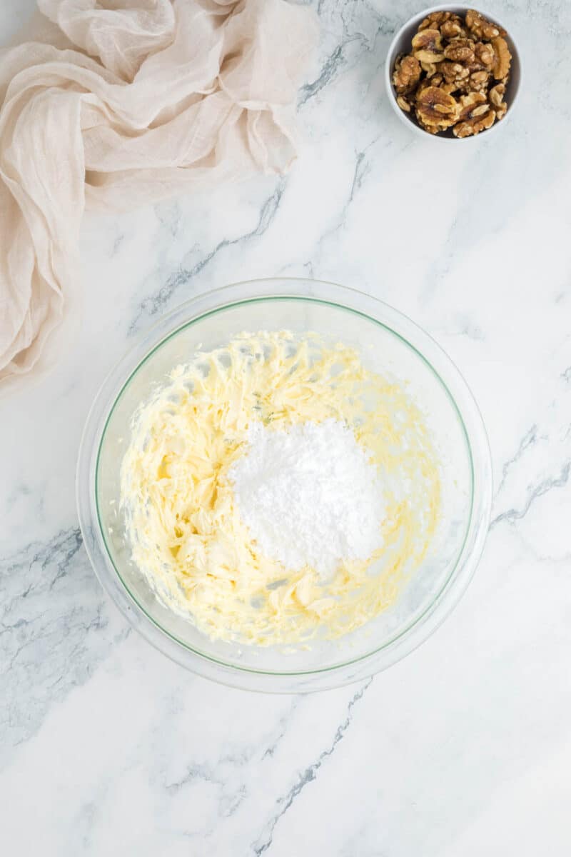 A glass bowl with creamed butter and powdered sugar on a marble countertop; a small bowl of walnuts is nearby.