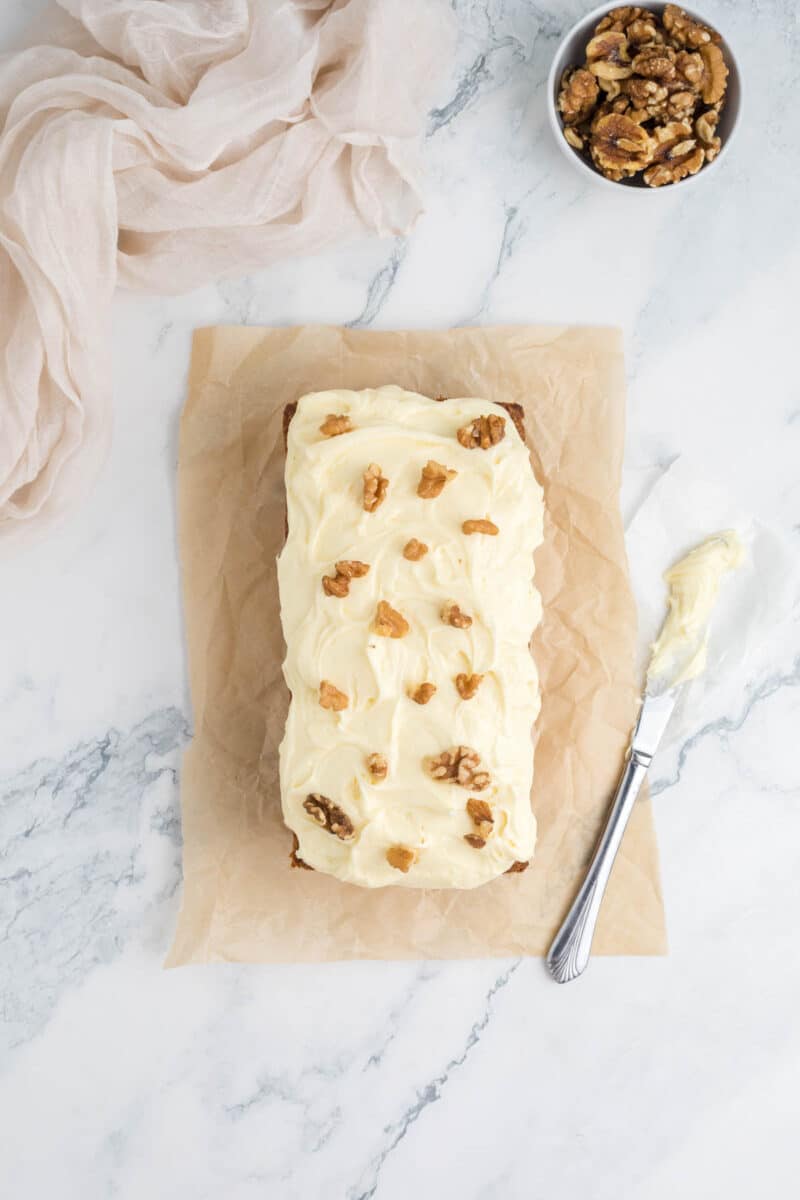 A loaf cake with white frosting and walnut halves on top sits on parchment paper, beside a bowl of walnuts and a knife with frosting on a marble surface.