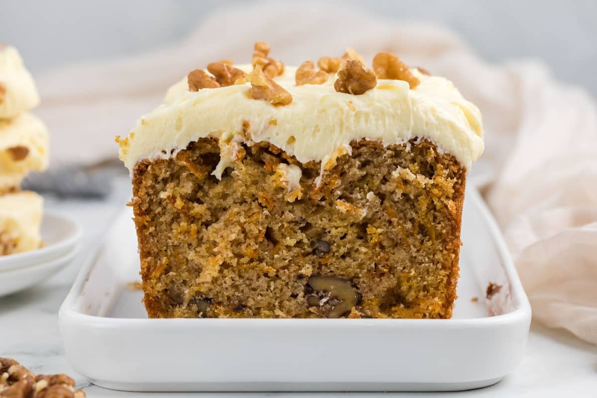 A sliced loaf of carrot cake with cream cheese frosting and walnut pieces on top, displayed on a white rectangular plate.
