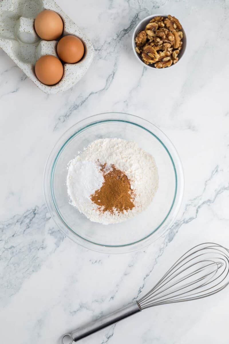 A glass bowl with flour, baking powder, and cinnamon on a marble surface, with eggs, walnuts, and a metal whisk nearby.