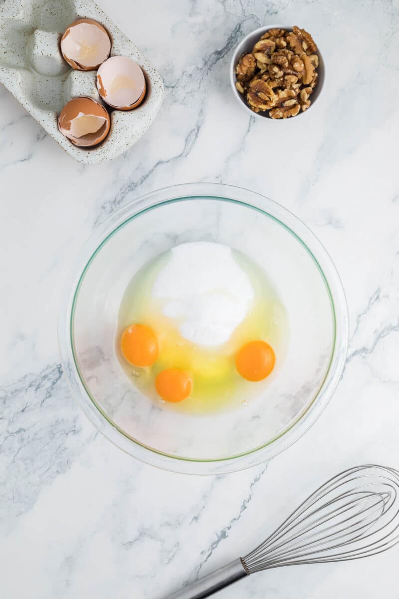 A glass bowl with three eggs and sugar, a whisk, a small bowl of walnuts, and cracked eggshells on a marble countertop.