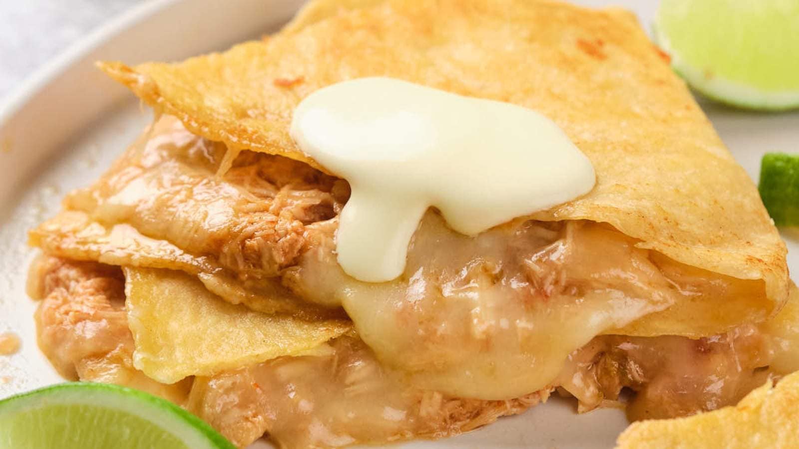 Close-up of a cheesy quesadilla filled with shredded chicken, topped with cream, and served with lime wedges on the side.