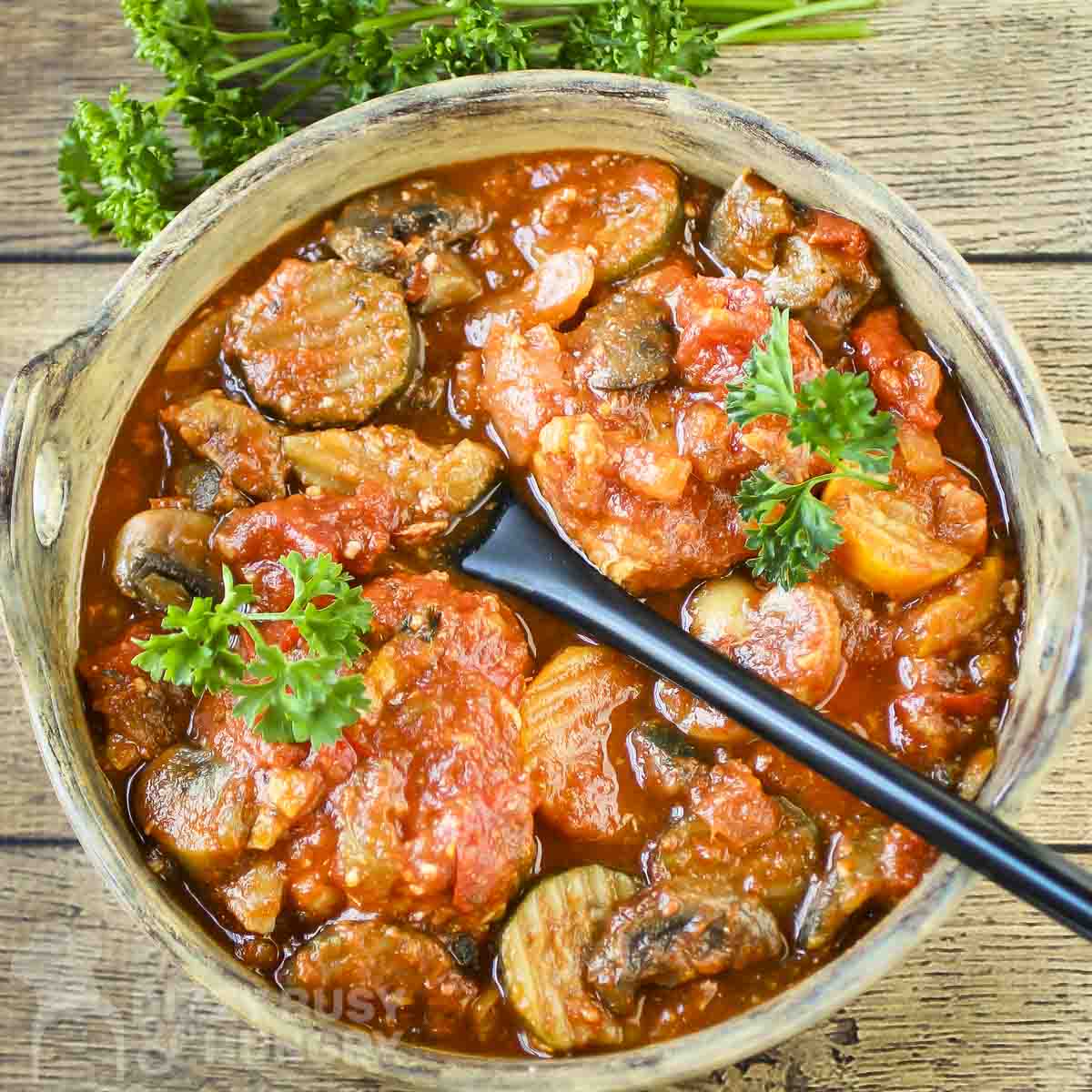 Overhead shot of chicken cacciatore in a large serving bowl with a black spoon inside on a wooden surface with a bunch of fresh parsley on the side.