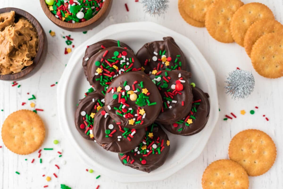 A plate of chocolate-covered cookies with colorful sprinkles, surrounded by crackers, a bowl of peanut butter, and small silver decorations on a white surface.
