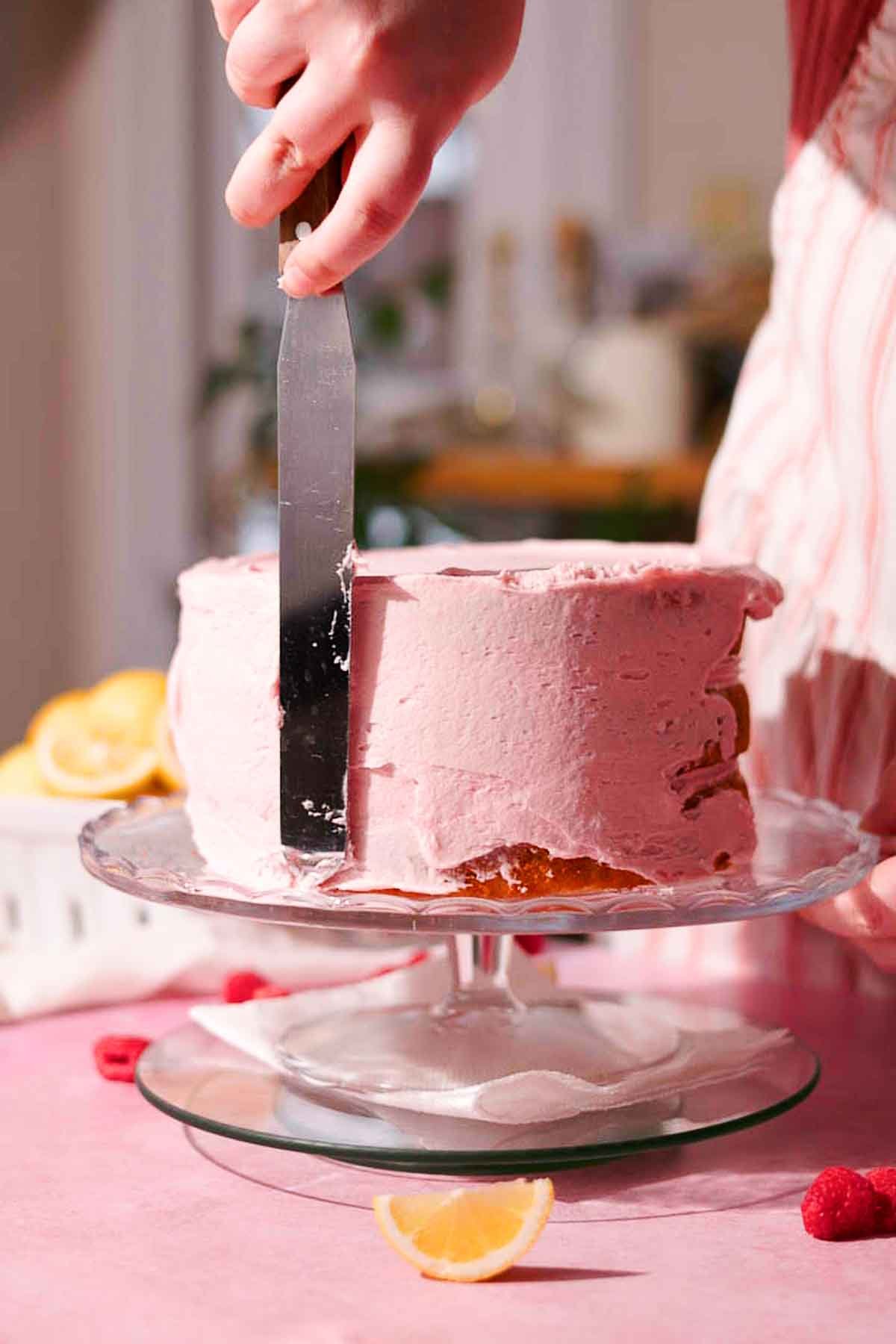 covering the sides of the cake with pink frosting using a long metal spatula 