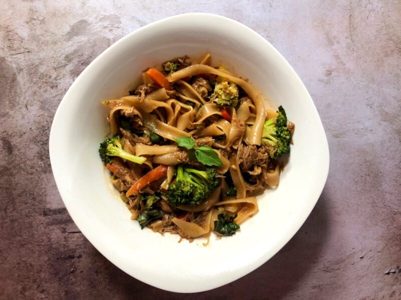 homemade pork drunken noodles in a white bowl with fresh vegetables and thai basil
