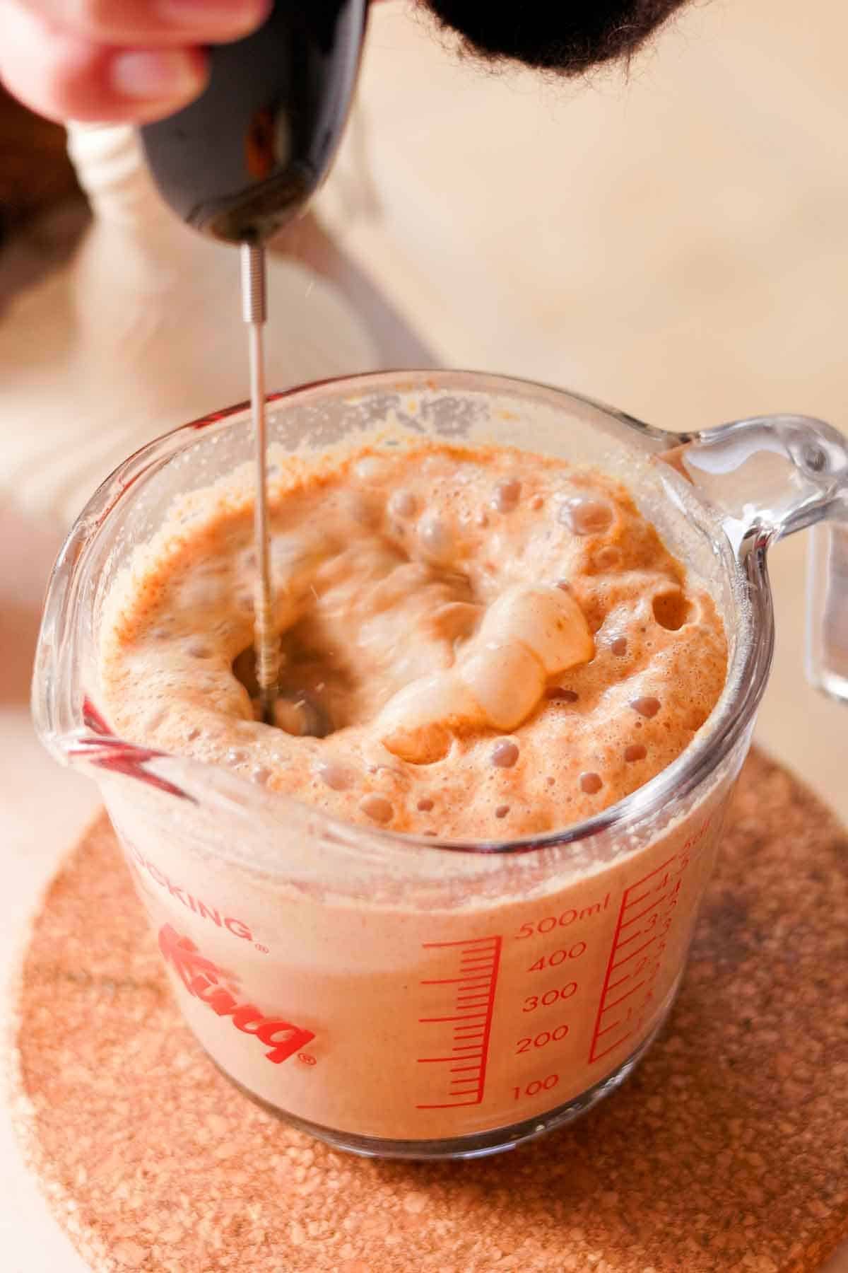 frothing pumpkin chai latte in glass measuring cup with electric whisk frother