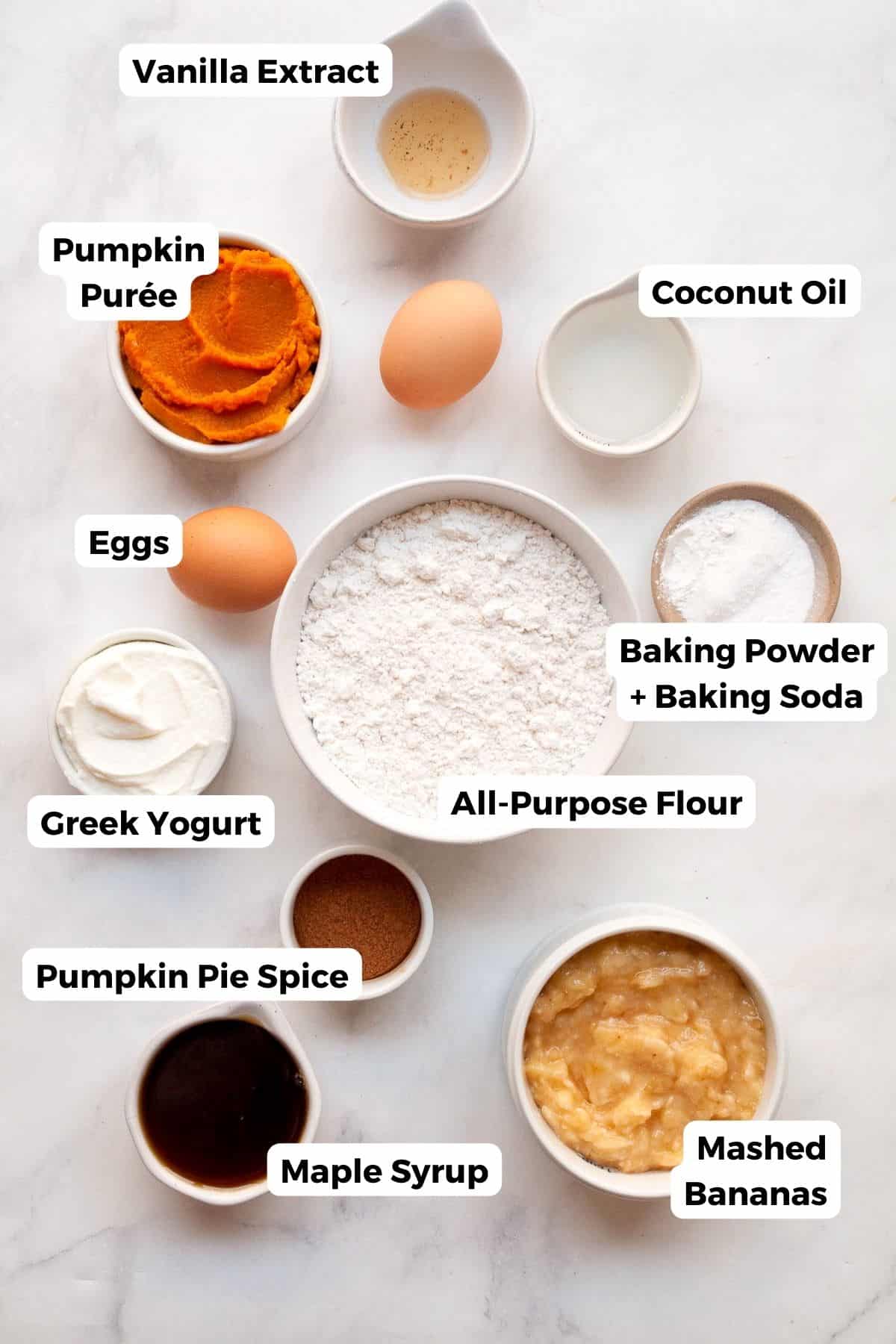 All the ingredients for healthy banana pumpkin muffins in small bowls with labels — ripe bananas, pumpkin purée, maple syrup, Greek yogurt, eggs, flour, baking powder, baking soda, pumpkin pie spice, cinnamon, and salt.