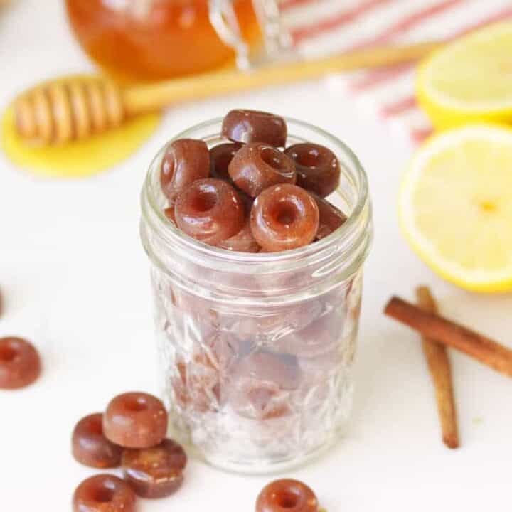 a mason jar of homemade cough drops with ginger root, honey on a honey stir stick, lemon and cinnamon sticks on the table next to it.