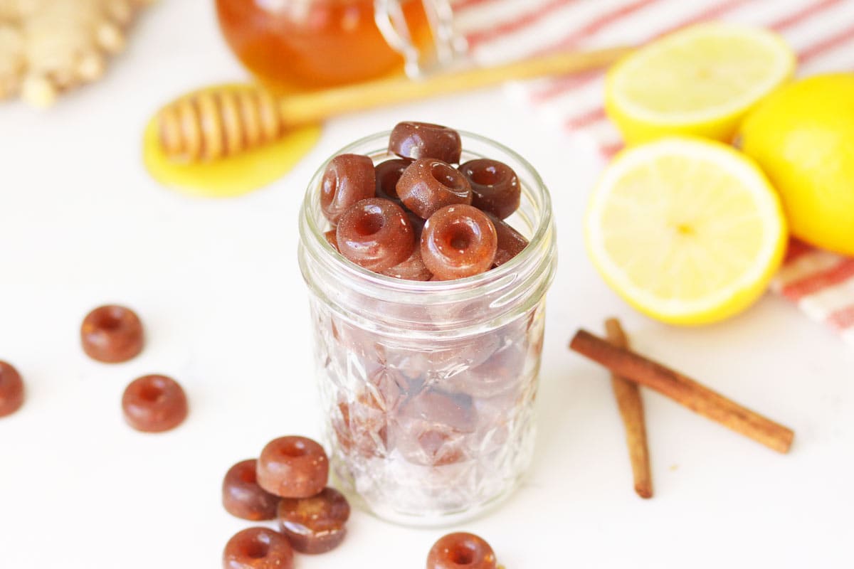 a mason jar of homemade cough drops with ginger root, honey on a honey stir stick, lemon and cinnamon sticks on the table next to it.
