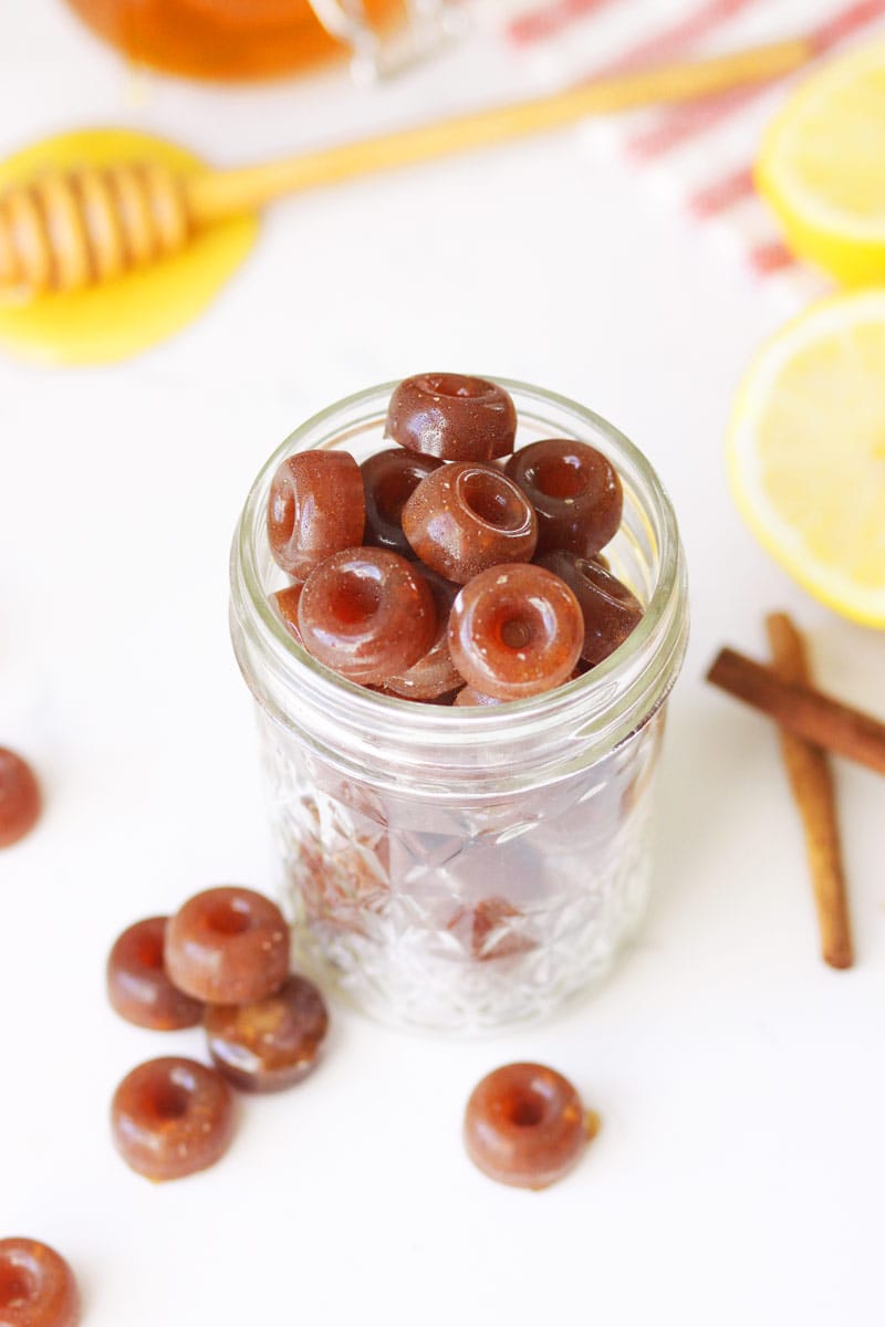 a mason jar of homemade cough drops with ginger root, honey on a honey stir stick, lemon and cinnamon sticks on the table next to it.