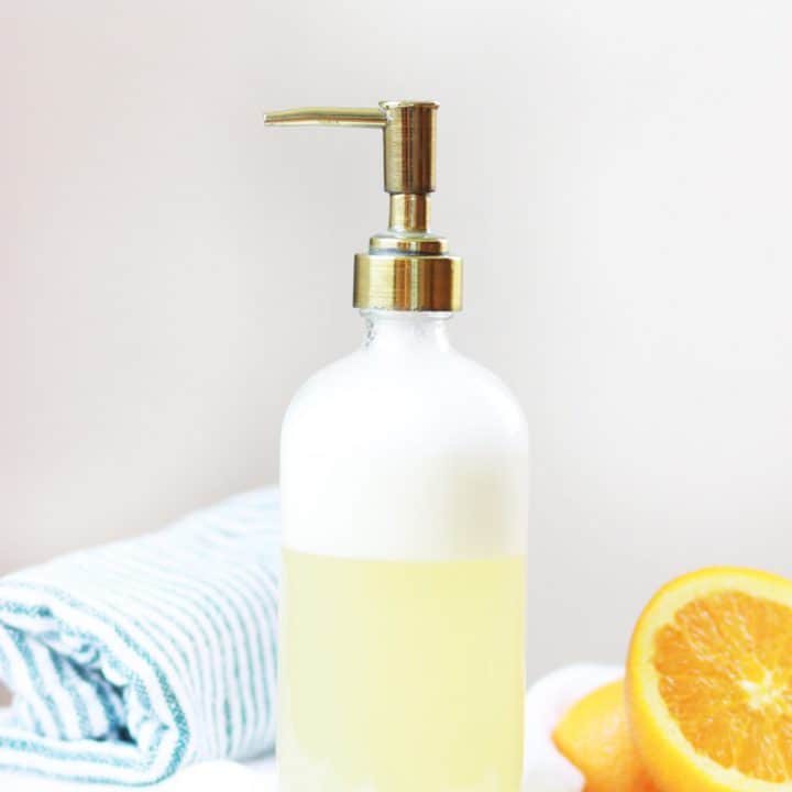 a glass bottle with face wash and a brass pump top. an orange and a striped bath towel are in the background