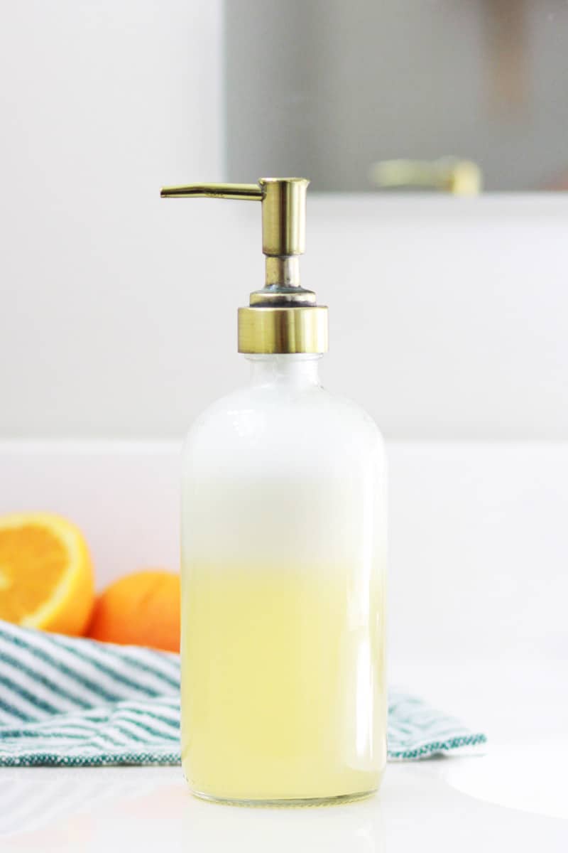 a glass bottle of homemade orange face soap with a cut orange and striped towel in the backgroung