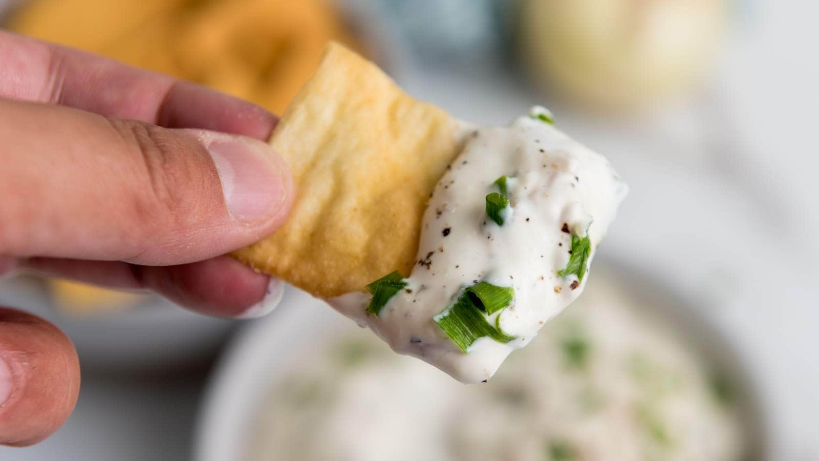 A hand holds a cracker dipped in a creamy onion dip topped with chopped chives.