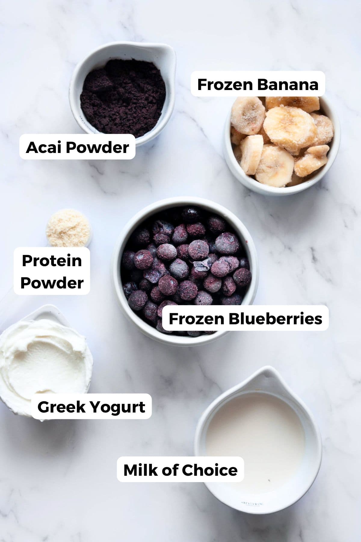 All ingredients in a homemade high-protein acai bowl labeled, including: frozen banana, mixed berries, Greek yogurt, vanilla protein powder, acai powder, and milk of choice.