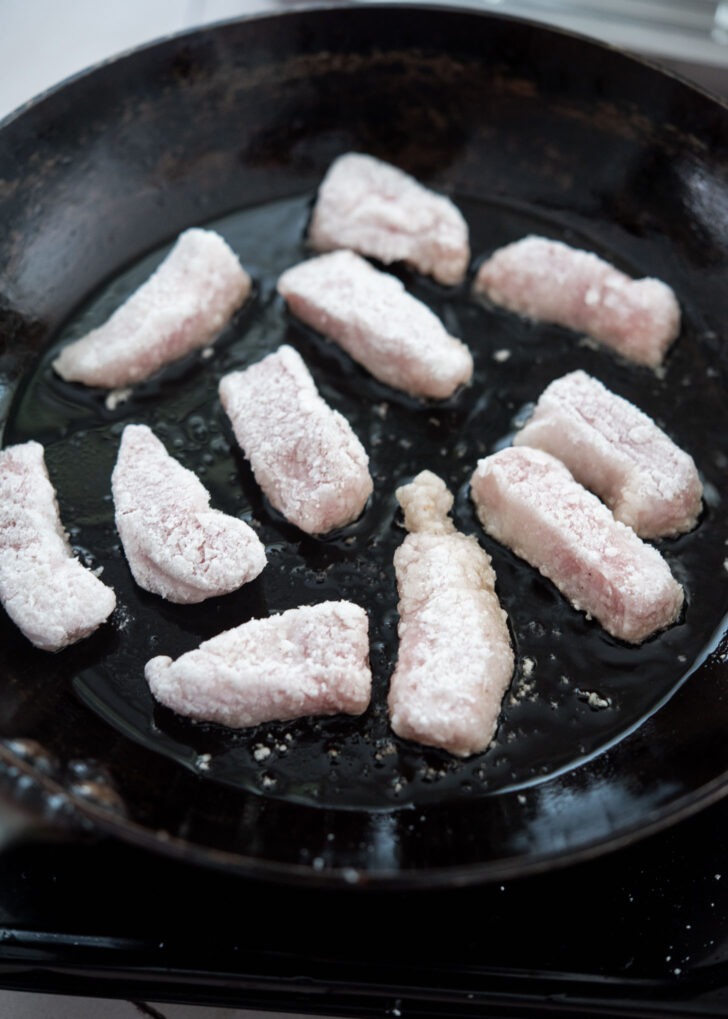 Starch-coated pork pieces being added to a hot skillet with oil for frying
