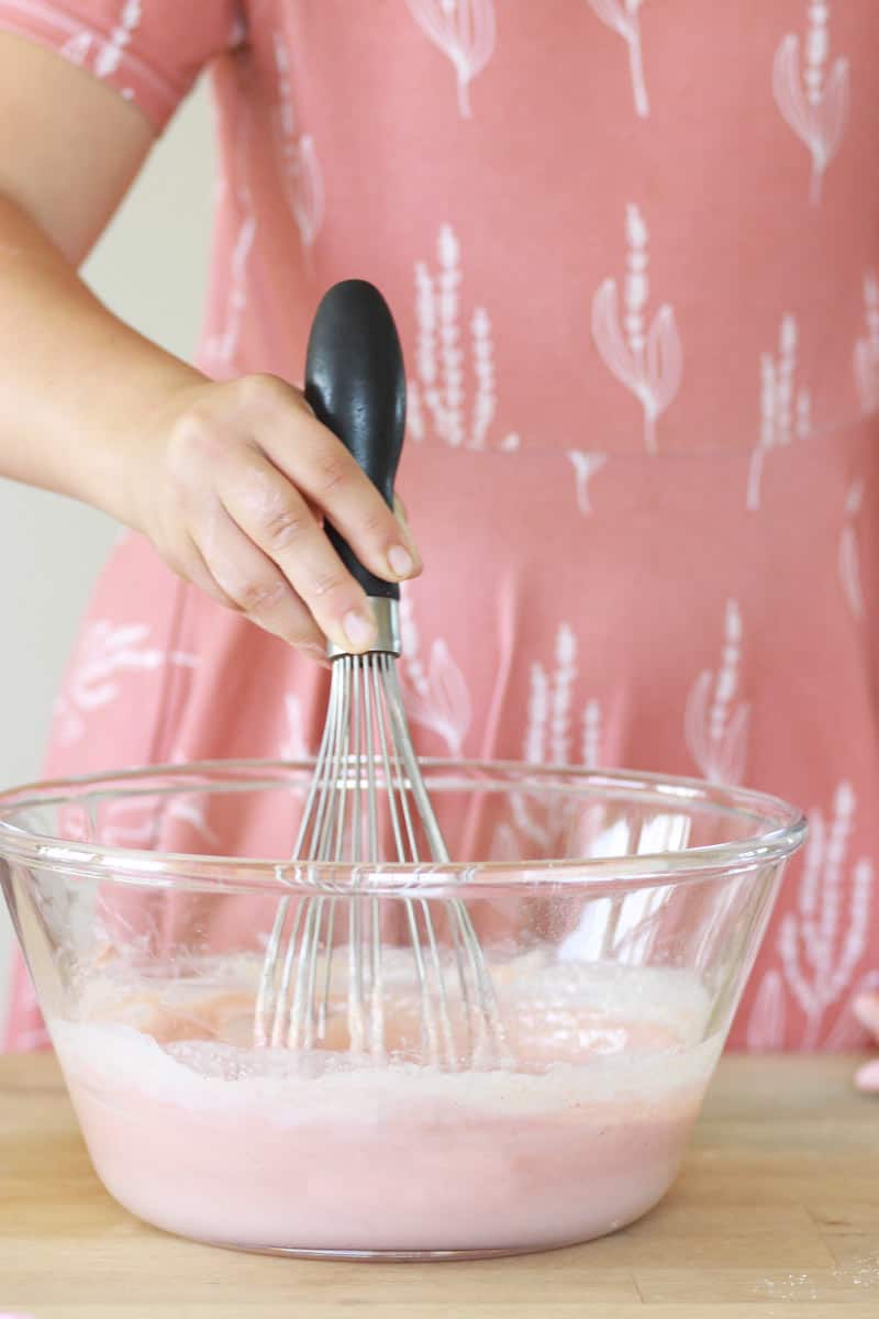 a girl in a pink dress whisks a bowl of melted soap mixture in a glass bowl