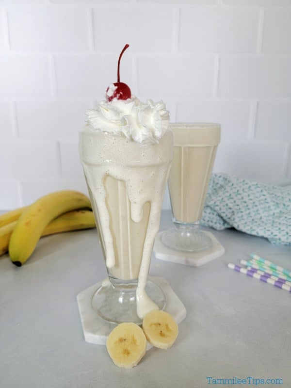 Two banana milkshakes in shake glasses with whipped cream and maraschino cherry garnish