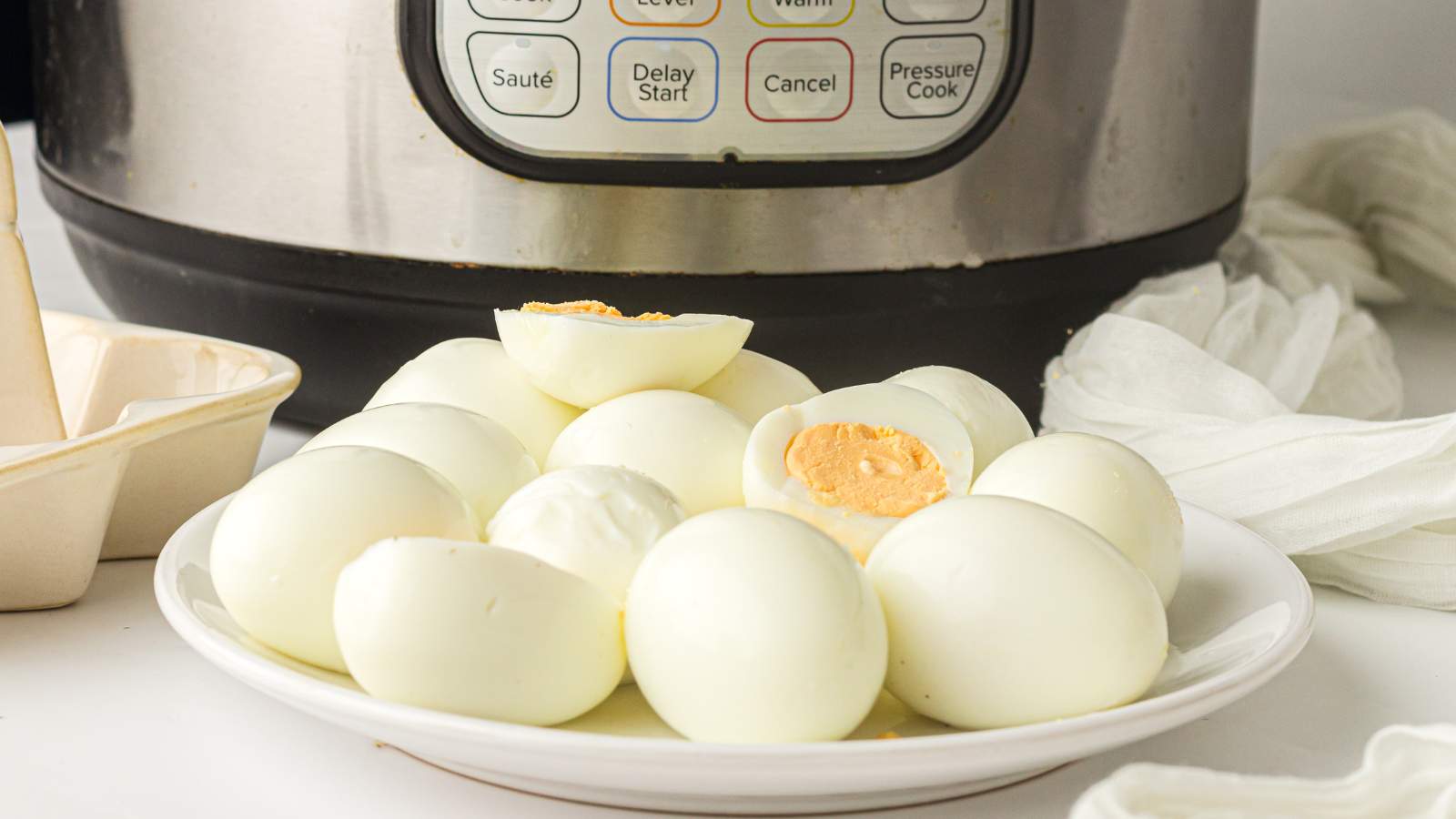 A plate of peeled hard-boiled eggs is placed in front of an instant pot on a white countertop.
