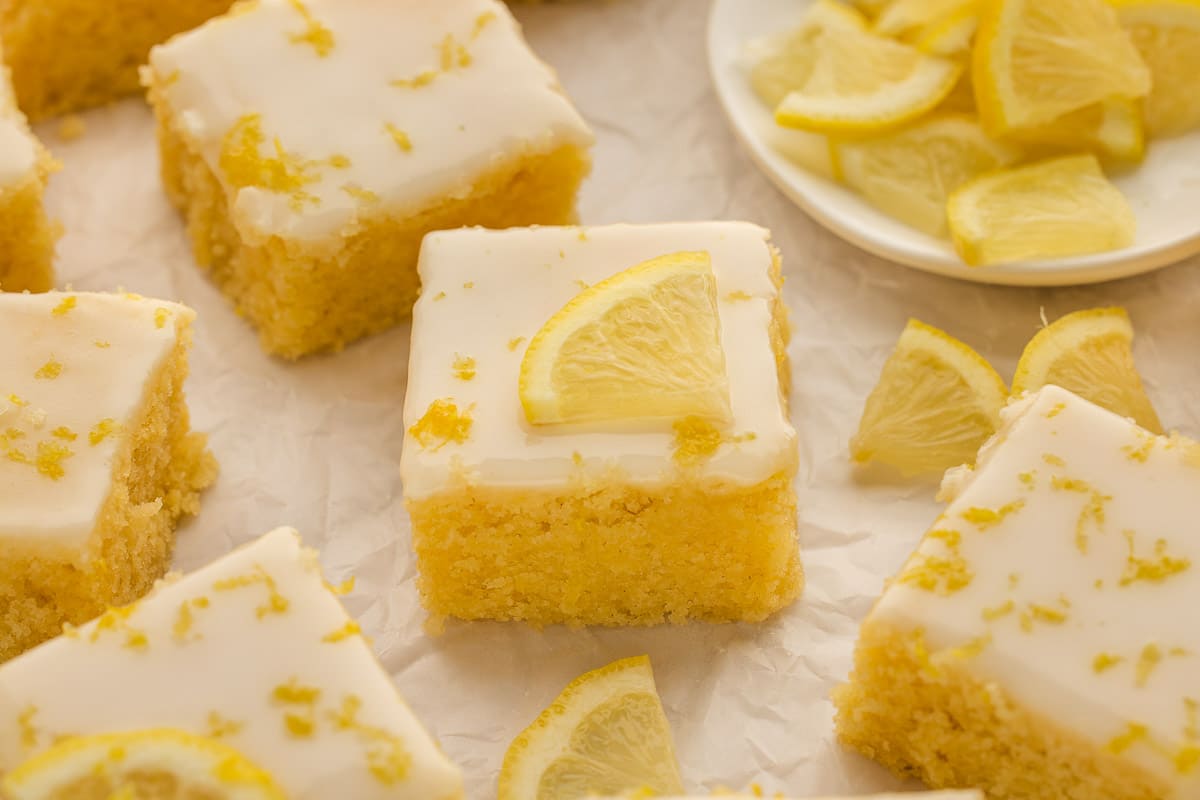 Lemon brownies with white icing, topped with lemon slices and zest, arranged on parchment paper with a plate of lemon slices in the background.
