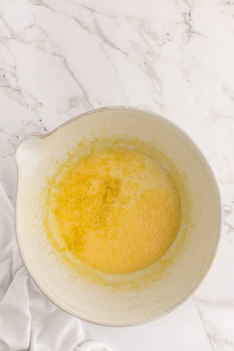 A mixing bowl containing a yellow, frothy liquid mixture sits on a white marble surface next to a white cloth.