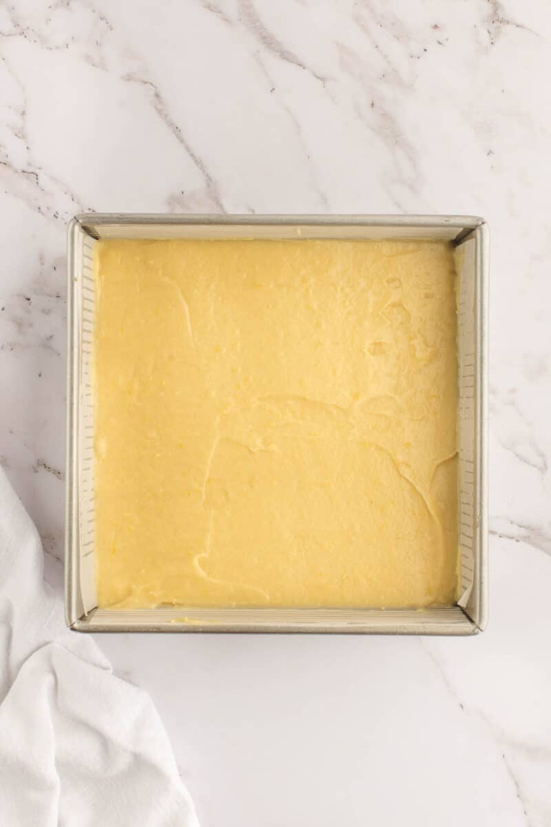 A square metal baking pan filled with smooth, unbaked yellow cake batter on a white marble surface.