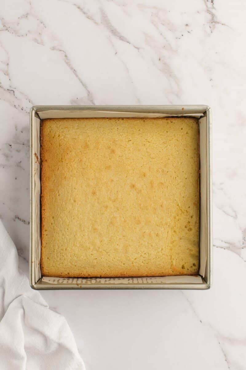 A square pan of plain, golden baked cake sits on a white marble surface with a white cloth partially visible at the bottom left.