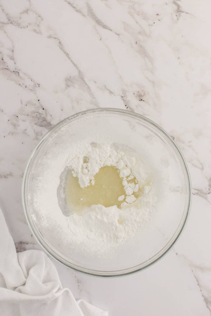 A glass bowl containing powdered sugar and a pool of liquid, set on a white marble surface with part of a white cloth visible.