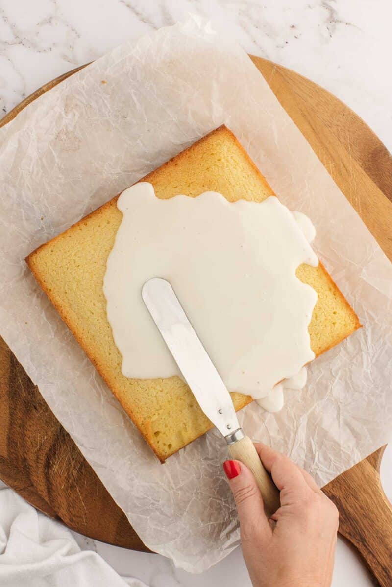 A hand spreads white icing onto a square sponge cake with a spatula, on parchment paper over a wooden board.