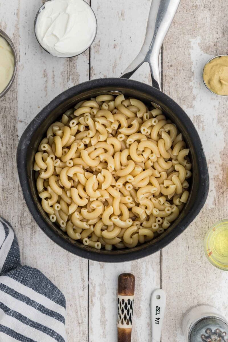 A black saucepan filled with cooked elbow macaroni sits on a white wooden surface surrounded by various small bowls and utensils.