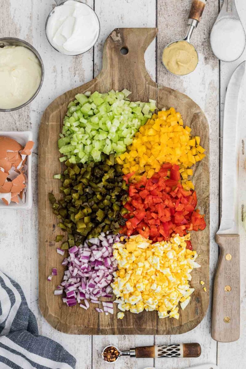 Chopped celery, yellow bell pepper, red bell pepper, pickles, red onion, and hard-boiled eggs arranged on a wooden cutting board with condiments nearby.