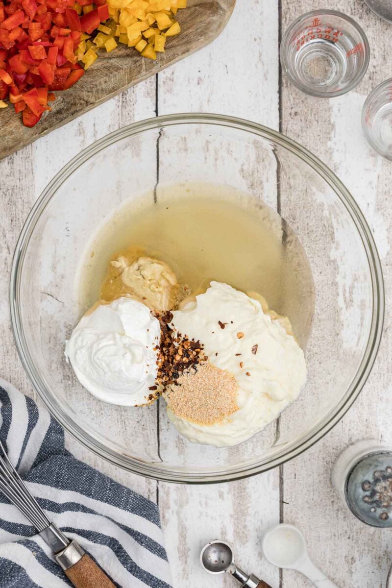A glass bowl with sour cream, mayonnaise, mustard, spices, and vinegar, next to chopped bell peppers on a wooden surface with measuring utensils nearby.