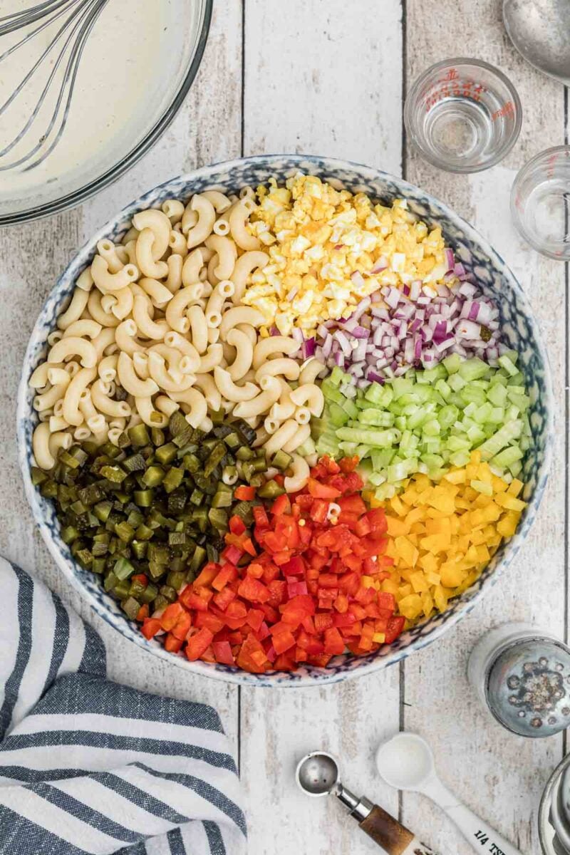 A large bowl containing separate sections of cooked macaroni, chopped hard-boiled egg, red onion, celery, yellow bell pepper, red bell pepper, and pickles.