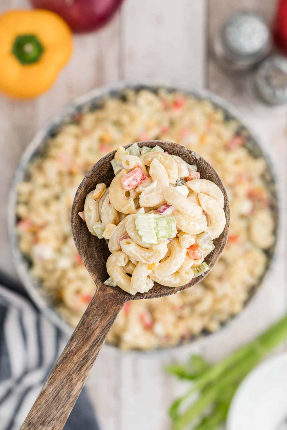 A wooden spoon holds Southern macaroni salad with chopped celery and red bell pepper above a bowl of the same dish on a wooden table.