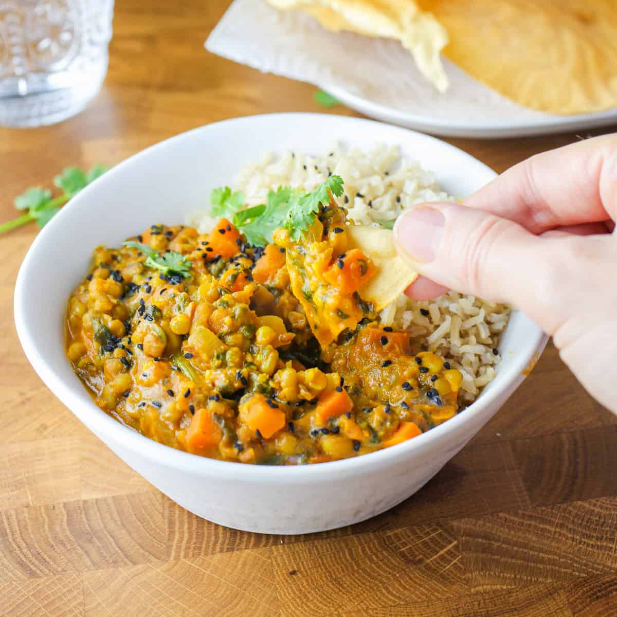 Photo showing a white bowl filled with mung bean curry and brown rice, with someone dipping a piece of poppadom into the curry, all on a wooden tabletop with poppadoms in the background.
