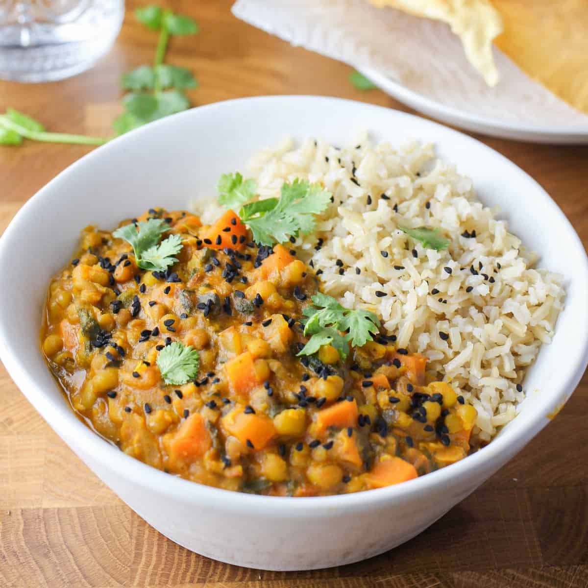 Photo showing a white bowl with Mung Bean Curry and brown rice, all set on a wooden table with a plate of poppadoms in the background and a glass of water and some coriander leaves sprinkled around.