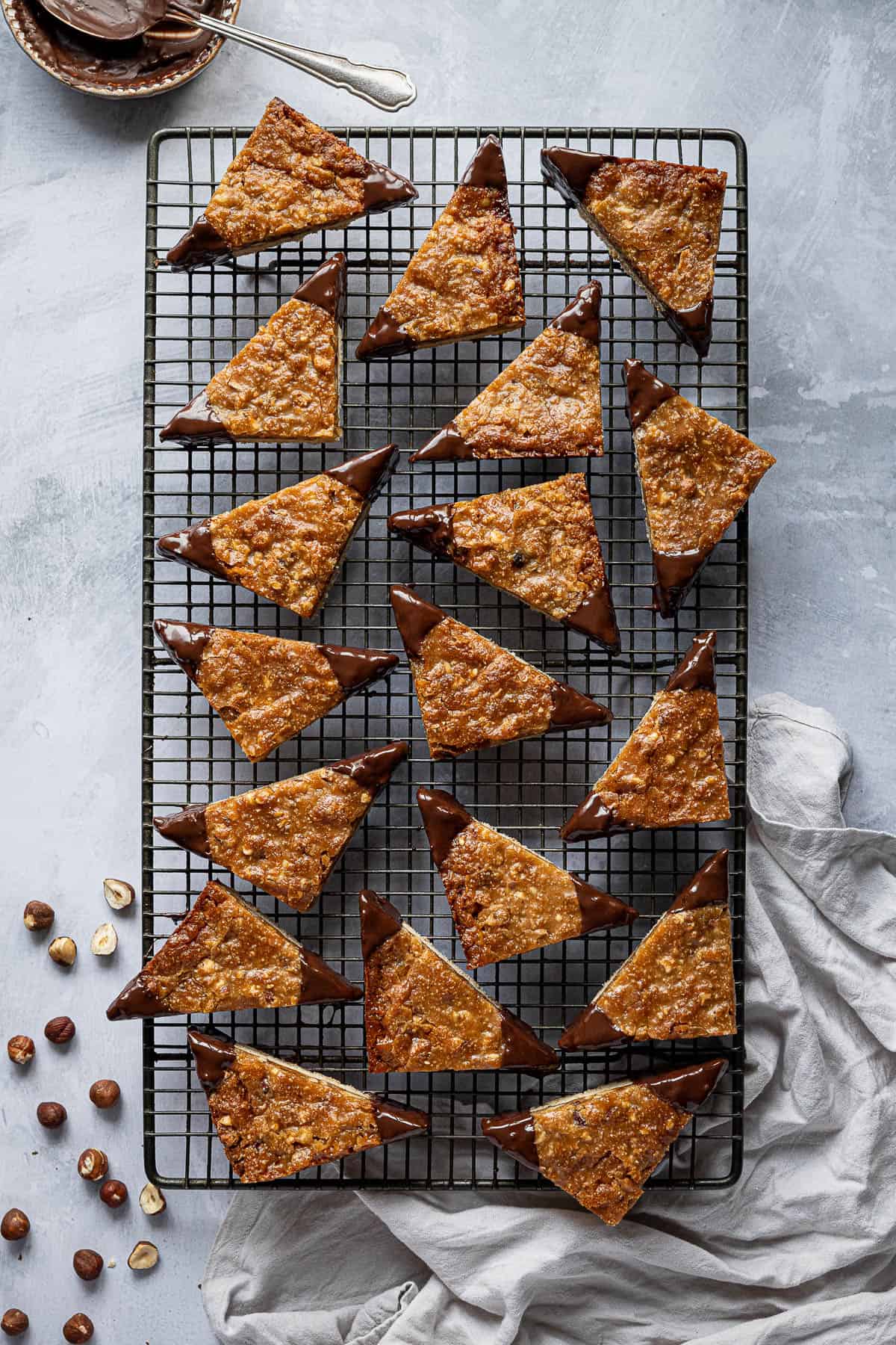 Vegan nussecken on a rectangular wire cooling rack with a bowl of chocolate, grey cloth and hazelnuts.