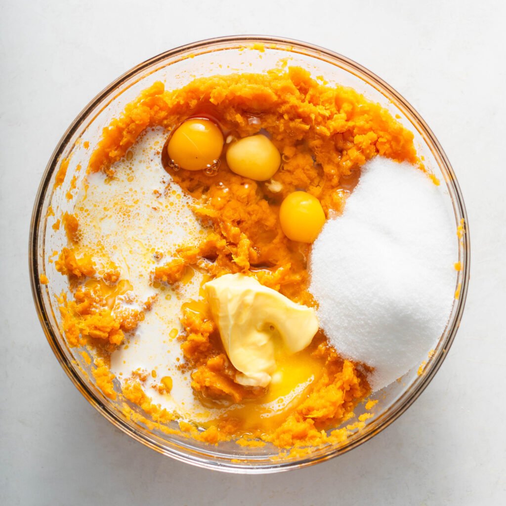 Mashed sweet potatoes, eggs, white sugar, melted butter, vanilla extract, and milk in a glass mixing bowl