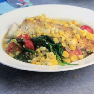 Oven baked fish in a white bowl with fresh summer vegetables like spinach, corn, and tomatoes will be one of your weeknight favorites! This recipe is quick and easy and everyone loves it!